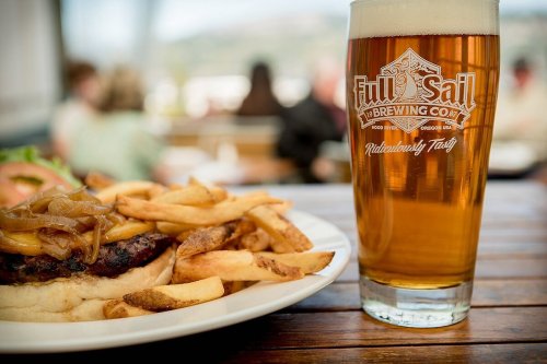 A plate of a burger with caramelized onions and fries next to a glass of amber beer on a wooden table.