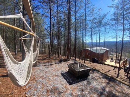 A serene outdoor space with hammocks, a fire pit, and a cabin surrounded by tall trees and mountains in the background.