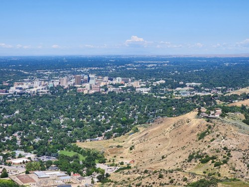 A panoramic view of a city skyline surrounded by green hills and open landscapes under a clear blue sky.