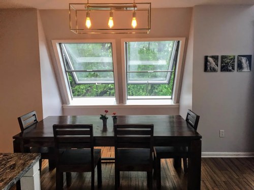 A dining area with a wooden table and chairs, featuring large windows that open to a green view outside.