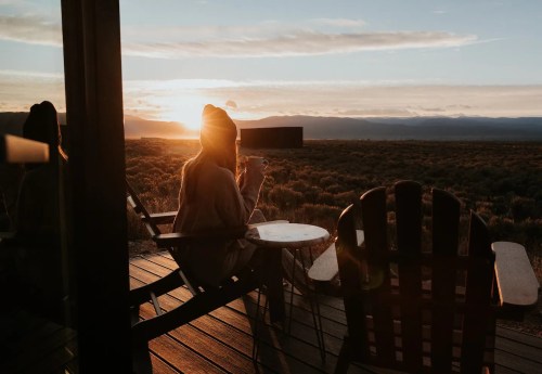 A person sits on a deck chair, enjoying a sunset over a vast landscape, with mountains in the distance.