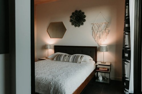 Cozy bedroom with a bed, decorative wall art, lamps, and a macramé hanging. Soft lighting creates a warm atmosphere.