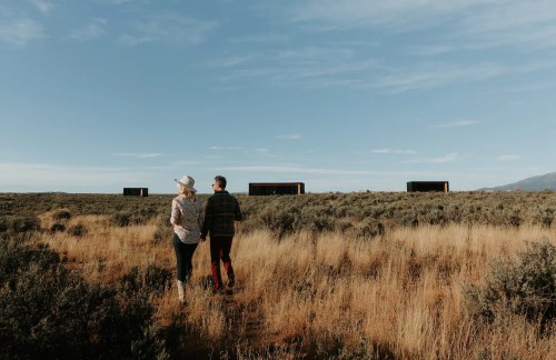 A couple walks hand in hand through a grassy landscape, with modern cabins visible in the distance under a clear sky.