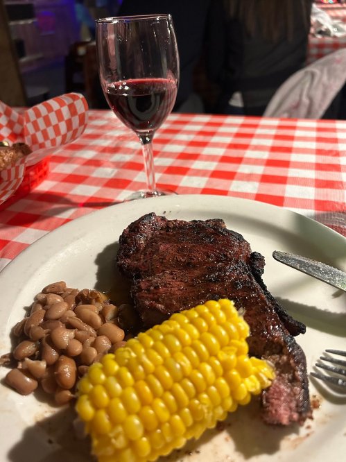 A plate with grilled steak, corn on the cob, and beans, accompanied by a glass of red wine on a checkered tablecloth.