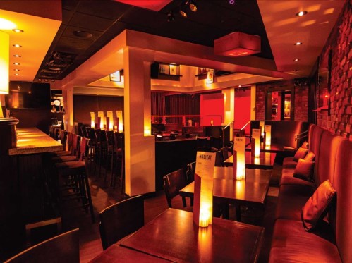 A dimly lit restaurant interior with wooden tables, chairs, and glowing candles, creating a warm, inviting atmosphere.