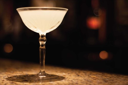 A elegant cocktail in a crystal glass sits on a bar counter, with a blurred background of warm lights.