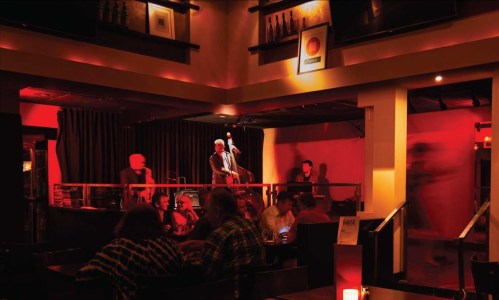 A dimly lit jazz club with a musician on stage, patrons enjoying drinks, and warm red lighting creating a cozy atmosphere.