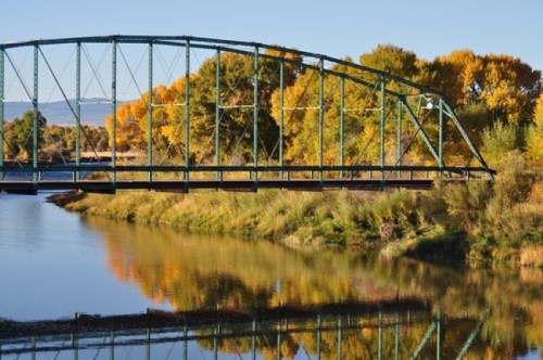 A green bridge spans a calm river, surrounded by trees with autumn foliage reflecting in the water.