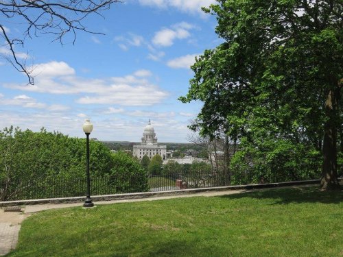 A view of a city skyline with a prominent dome building, framed by green trees and a blue sky with clouds.