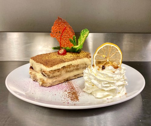 A slice of tiramisu topped with a strawberry, mint, and a cherry, served with a dollop of whipped cream and a lemon slice.