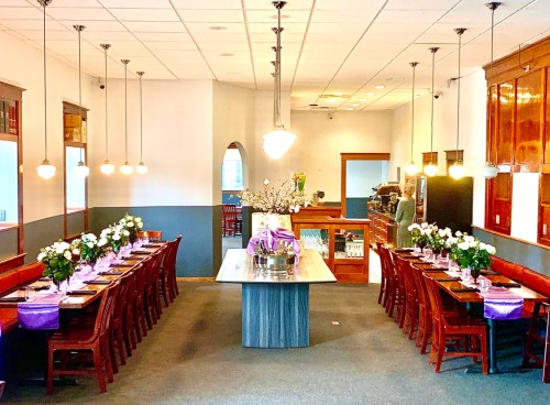 A spacious restaurant with a long table set for dining, adorned with flowers and purple accents, and warm lighting.