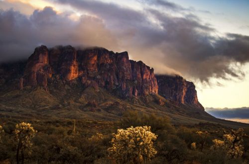 A dramatic mountain range under cloudy skies, with desert vegetation in the foreground.
