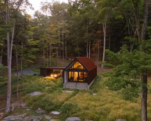 A modern cabin nestled in a forest, surrounded by greenery and rocks, with warm light glowing from its windows at dusk.