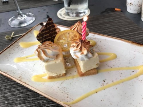 A dessert plate featuring a birthday cake slice with meringue, a candle, and lemon garnish on a decorative sauce.