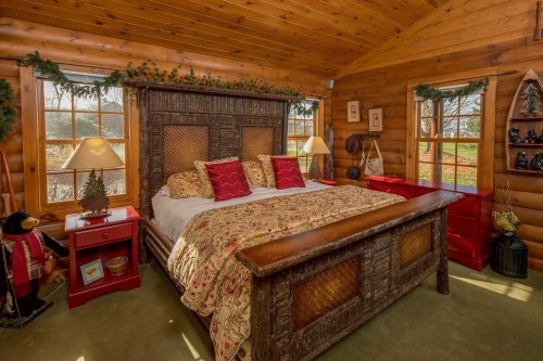 Cozy log cabin bedroom with a wooden bed, decorative pillows, red nightstands, and large windows overlooking greenery.
