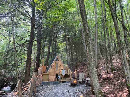 A cozy wooden cabin nestled in a forest, surrounded by trees and a small fire pit nearby.
