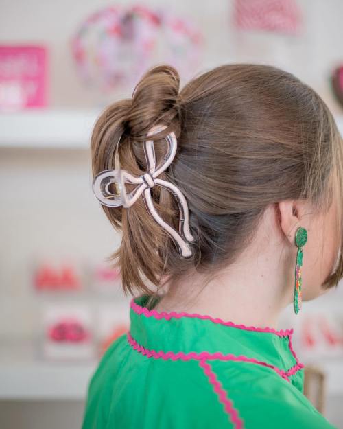 A woman with a stylish updo featuring a large bow hair clip, wearing colorful earrings and a green outfit.