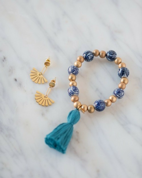 A blue and gold beaded bracelet with a tassel next to gold fan-shaped earrings on a marble surface.