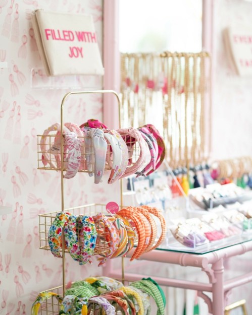 A display of colorful headbands on a gold rack, with a pastel pink wall and jewelry in the background.