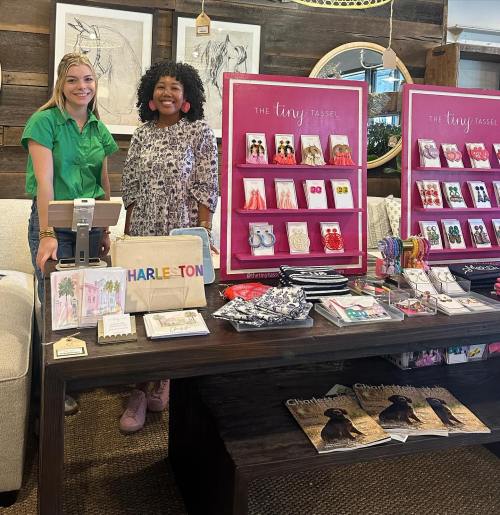 Two women stand behind a table displaying colorful accessories and products, with a pink backdrop featuring "The Tiny Tassel."