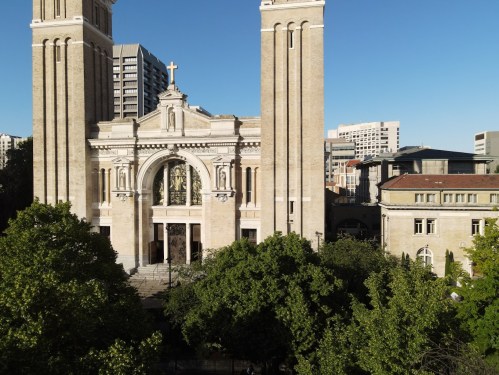 A large church with a cross on top, surrounded by trees and modern buildings in the background under a clear blue sky.