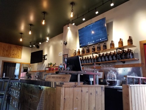 A cozy bar counter with wooden accents, shelves of growlers, and two TVs mounted on the wall. Warm lighting overhead.