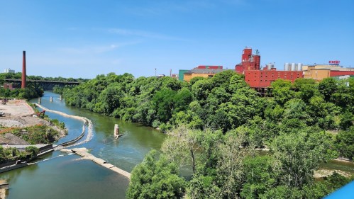 A river flows through lush greenery, with industrial buildings visible in the background under a clear blue sky.