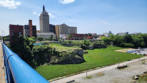 Cityscape featuring buildings, green space, and a clear blue sky, viewed from a bridge.