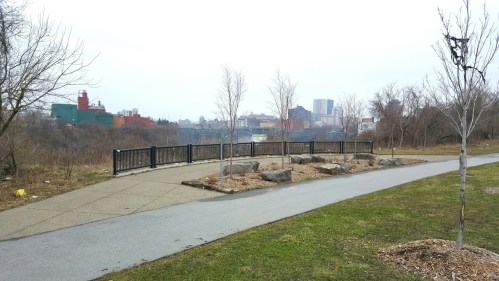 A park path with a railing, bare trees, and a view of buildings in the distance on a cloudy day.