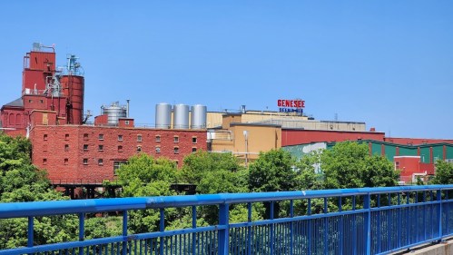 View of the Genesee Brewery with red brick buildings and silos, surrounded by greenery under a clear blue sky.