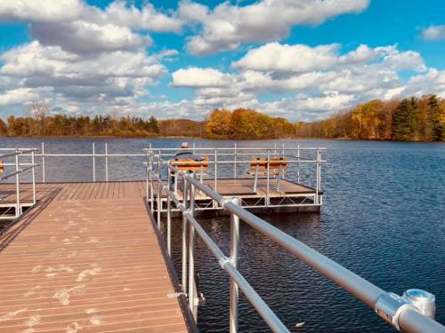 A wooden dock extends over a calm lake, with a person sitting on a bench, surrounded by trees and fluffy clouds.