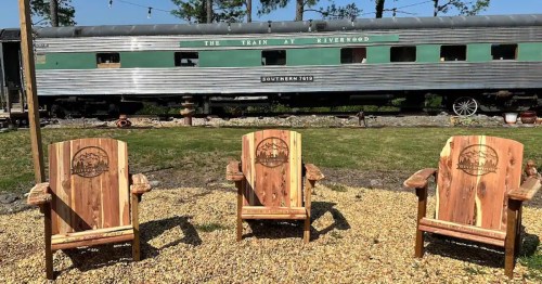 Three wooden chairs in front of a vintage train car, set in a grassy area with trees in the background.