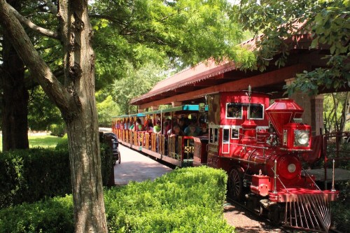 A red train with passengers is parked under a shaded area, surrounded by greenery and trees.