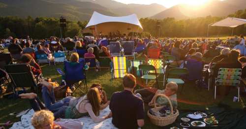A crowd enjoys a concert outdoors at sunset, with people sitting on blankets and chairs in a scenic mountain setting.