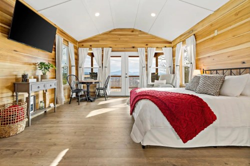Cozy cabin interior with wooden walls, a bed with a red throw, a small dining table, and large windows showcasing a view.