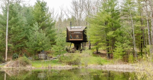 A modern treehouse-style cabin stands on stilts by a serene pond, surrounded by lush green trees.