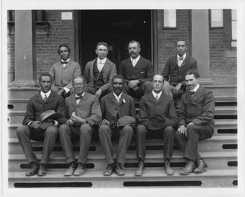 A historic black-and-white photo of nine men seated on steps, dressed in formal attire from the early 20th century.