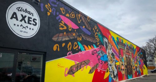Colorful mural on a building featuring abstract shapes, feathers, and vibrant patterns, with "Urban Axes" logo.