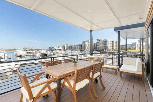 A spacious balcony with a wooden dining table overlooks a marina filled with boats and modern buildings in the background.