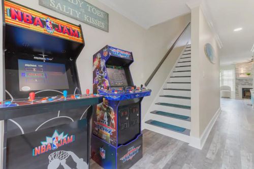 Two arcade machines, NBA Jam and NFL Blitz, are positioned near a staircase in a bright, modern hallway.
