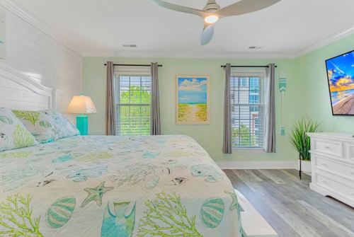 Bright bedroom with a large bed, light green walls, and windows, featuring beach-themed decor and a mounted TV.