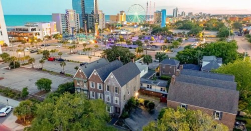 Aerial view of a coastal city with a ferris wheel, modern buildings, and a mix of greenery and parking lots.