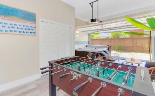 A foosball table in a bright, open space with a garage door, art on the wall, and a hot tub in the background.