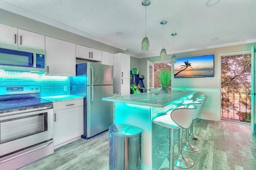 Modern kitchen with sleek appliances, glowing blue accents, and a view of a sunset through a large window.