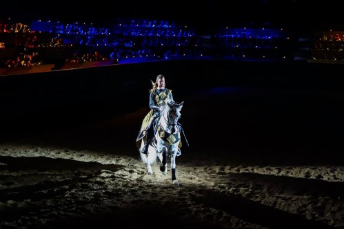 A knight in armor rides a white horse in a dimly lit arena, showcasing a dramatic performance.