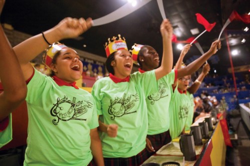 A group of excited students in green shirts and crowns cheer enthusiastically at an event, waving ribbons and flags.