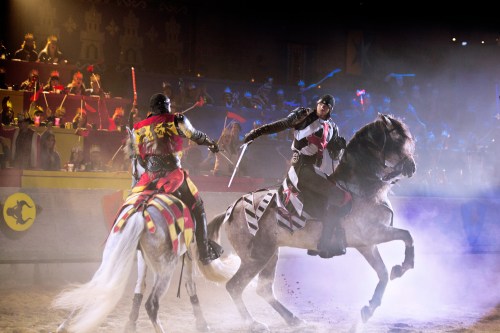 Two knights in armor duel on horseback in a medieval arena, surrounded by a cheering crowd. Dust and smoke fill the air.