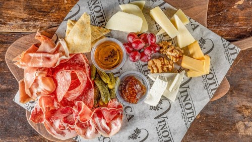 A wooden platter with assorted meats, cheeses, grapes, nuts, pickles, and honey, arranged on patterned paper.