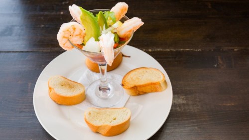 A shrimp cocktail served in a glass, garnished with vegetables, accompanied by slices of toasted bread on a plate.