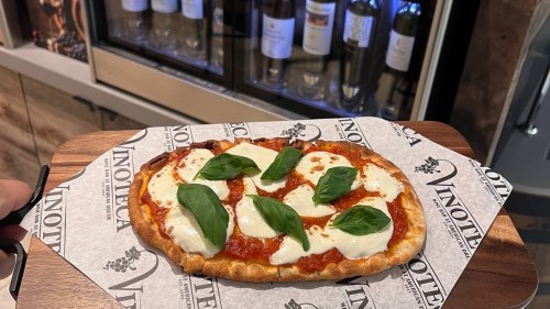 A hand holding a wooden board with a margherita pizza topped with fresh basil on a Vinoteca-branded paper.